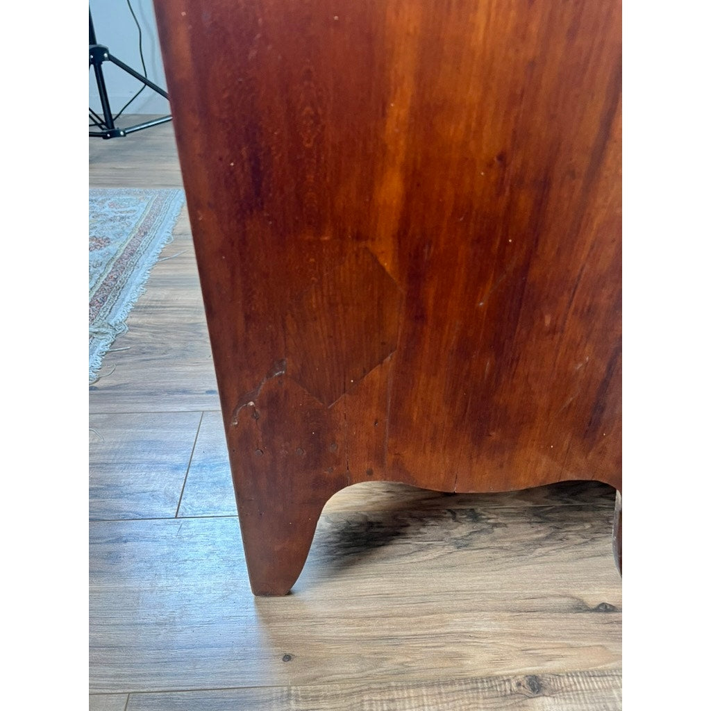 Close-up of a wooden furniture leg from a Rare Labeled William Lloyd Springfield Massachusetts Federal Cherry Chest of Drawers, circa 1800, showing a visible scratch and chipped area near the bottom; the piece is placed on a wood floor with a patterned rug partially visible in the background.