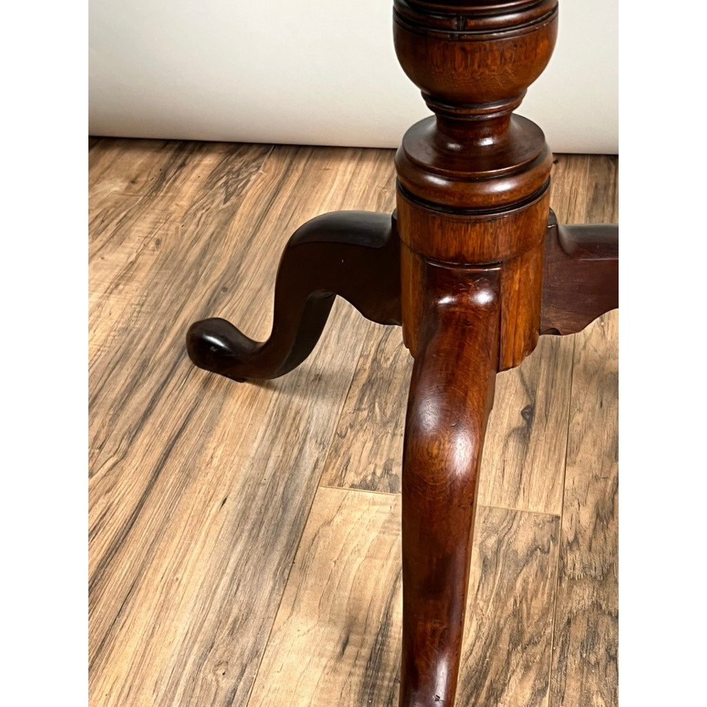 Close-up of a polished Antique Queen Anne Mahogany Tilt Top Breakfast Table leg, c. late 18th century, showing ornate curved detailing and fine craftsmanship as it rests on a wooden floor with contrasting grain patterns. Brand: Unknown.
