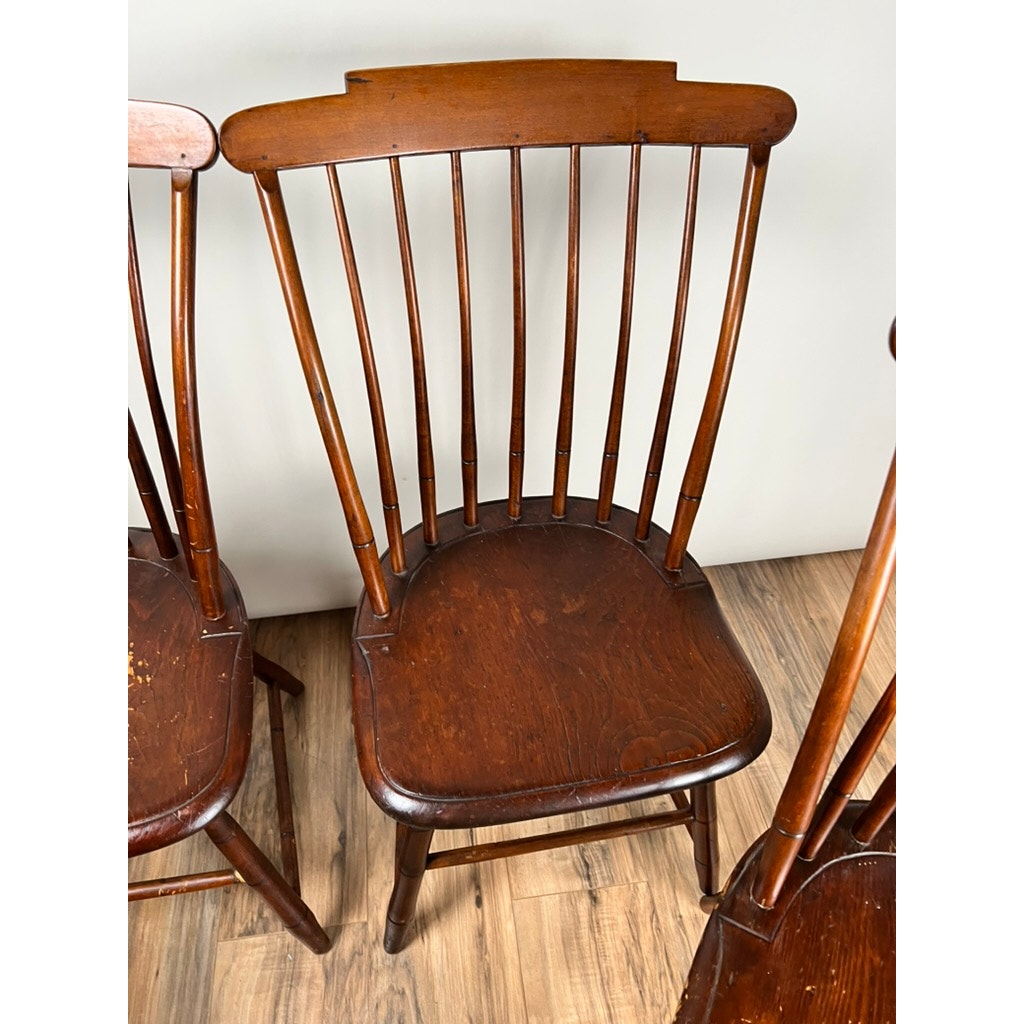 An Antique Set of Four New Hampshire Step Down Windsor Chairs by JR Hunt, c. Early 19th Century, features dark brown wood, curved backrests with vertical spindles, and contoured seats. Parts of other matching chairs appear on each side.