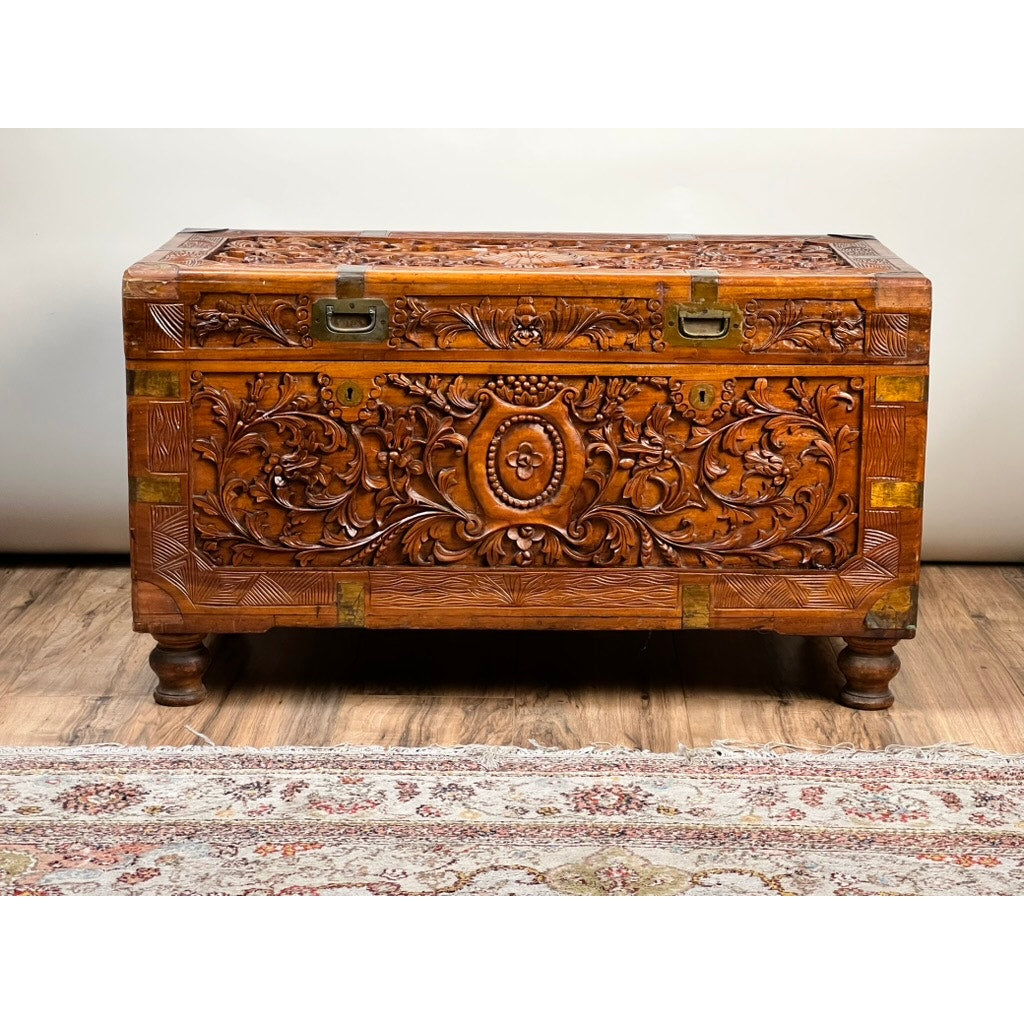 The Chinese Export Carved Camphor Wood Dowry Chest, featuring intricate floral carvings, metal latches, and short rounded legs, is placed on a wooden floor beside a patterned rug.