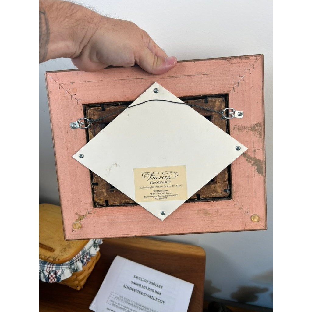 A hand holds the back of an antique pink-framed oil on canvas, "Landscape with Windmill," signed “Brüguer” (circa 1880-1910). The frame rests on a wooden surface with papers and cloth below; backing reads “Pierces Framing.”.