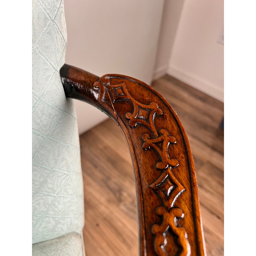Close-up of an Antique George II Mahogany Gainsborough Open Arm Chair (mid-19th century) shows its ornate carved frame, detailed armrest, and light diamond-patterned upholstery. Cabriole legs rest on a wooden floor against a white wall.