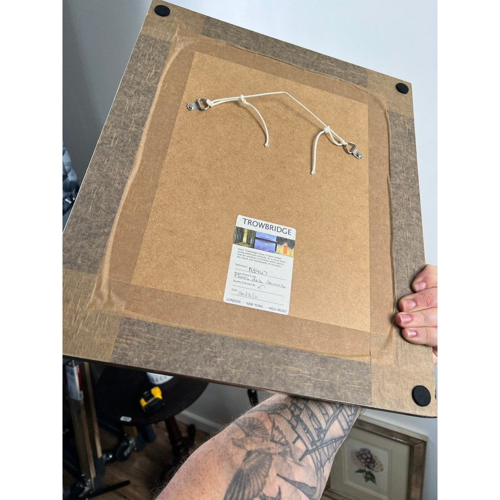 A tattooed arm holds up a Trowbridge Vintage Custom Framed Botanical Print “Campanula Vidalii” after Louis van Houtte (1810–1876), showing its cord and label, with part of a Hollywood Regency room and decor in the background.
