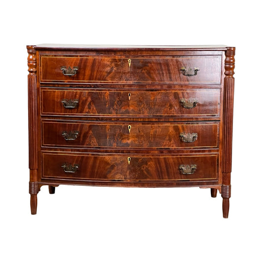 Antique Sheraton Mahogany Bowfront Chest, early 19th century, features four curved drawers with ornate brass handles and side carvings. Its polished mahogany finish highlights classic Sheraton style and craftsmanship. Brand: None/Unknown.