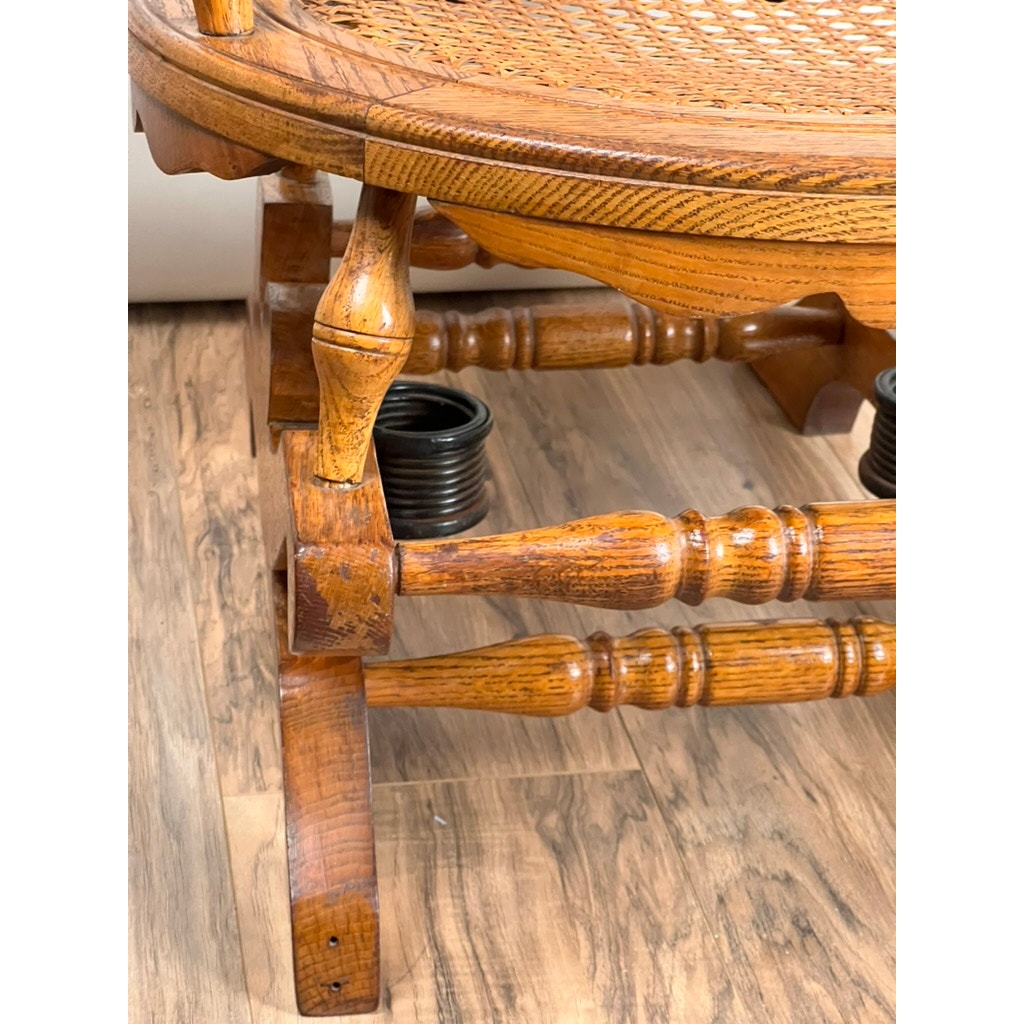 Close-up of the wooden leg and woven cane seat of an Antique Victorian Oak Platform Rocker (late 19th century), featuring decorative turned wood details. Metal springs for support are visible beneath, all resting on a wood floor. Brand unknown.