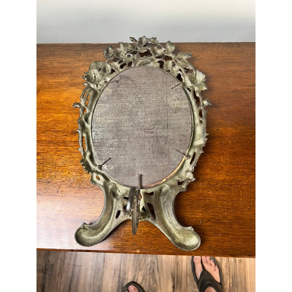 An Antique Art Nouveau Silvered Cast Brass Rose Vanity Dressing Mirror, dated 1909, rests on a wooden surface. Its ornate, leaf-like border recalls vintage elegance. A person in sandals is partly visible at the bottom of the image. Brand: Unknown.