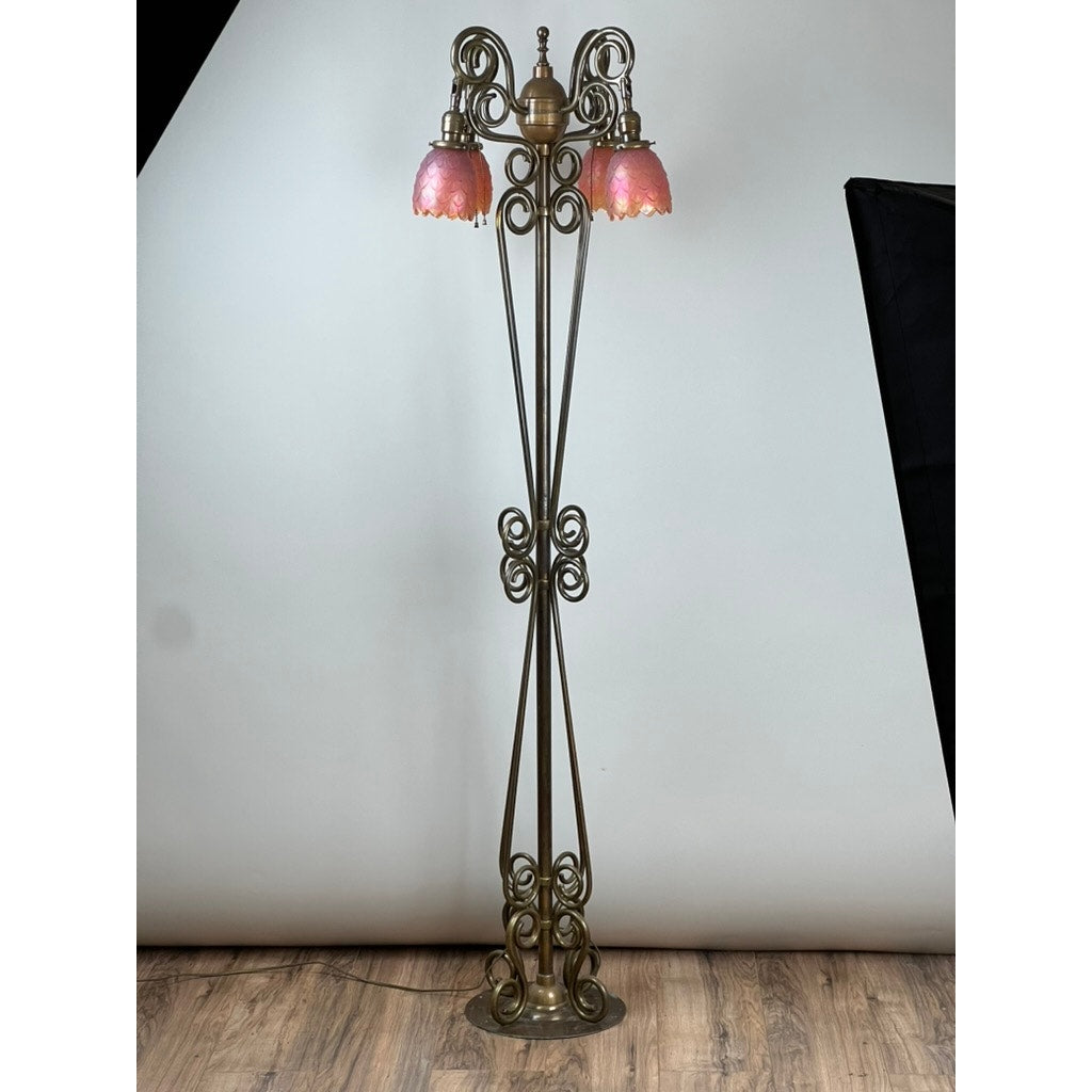 An Antique Art Nouveau Brass Floor Lamp, attributed to Jules Cayette and Charles Schneider (c. 1920), features ornate scrollwork and three upward-facing pink glass shades, standing on a wooden floor against a light backdrop.