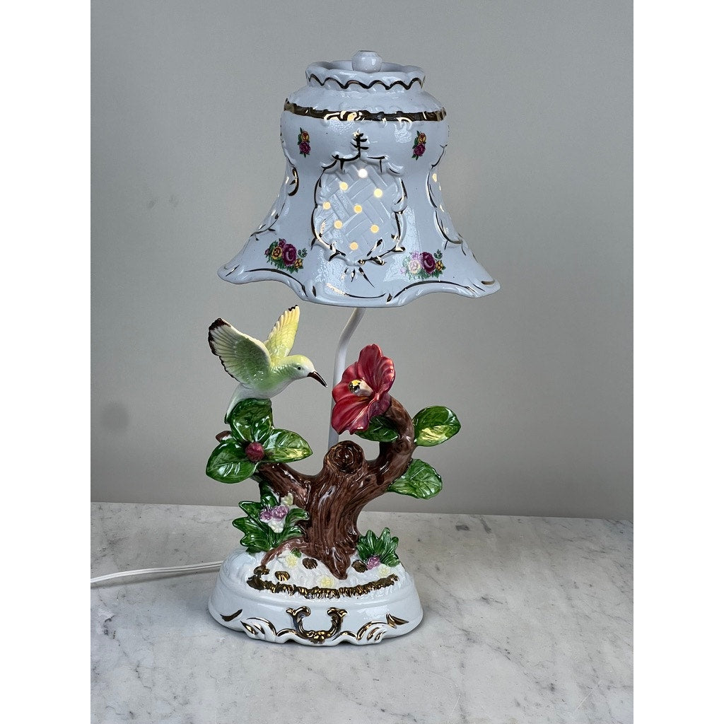 The Vintage Lena Liu Porcelain Hummingbird Accent Lamp features a floral-painted shade and a detailed base with a tree, red flower, green leaves, and yellow bird in flight, displayed on a gray marble surface.