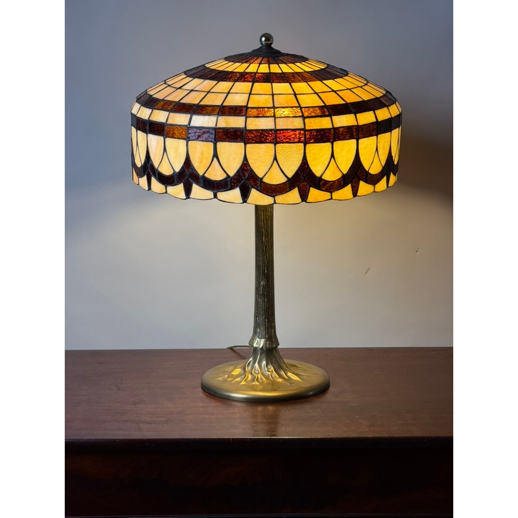 A Vintage Tiffany Revival Leaded Stained Glass Table Lamp with Cast Brass Tree-Form Base by Pearl City Antiques, featuring a yellow and brown stained glass shade and an intricate brass base, sits elegantly on a wooden surface against a plain background.