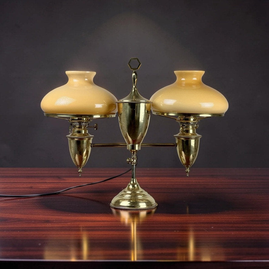An Antique Bradley and Hubbard Brass Double-arm Student Lamp with caramel shades by Bradley and Hubbard rests on a polished wooden table against a dark background.