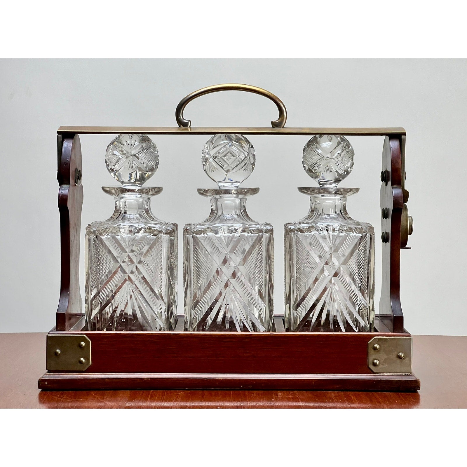 An Antique English Edwardian Betjemann’s Cut Crystal Three Bottle Tantalus by Betjemann's, c. early 20th century, displays three matching crystal decanters in an elegant holder on a wooden surface against a plain background.