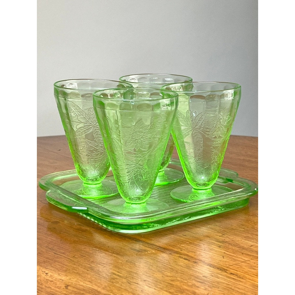 An Antique Jeannette Glass Co. Green Uranium Floral Poinsettia 5-piece beverage set from the 1930s, featuring four cups and a matching rectangular tray, is displayed on a wooden surface against a plain light background.