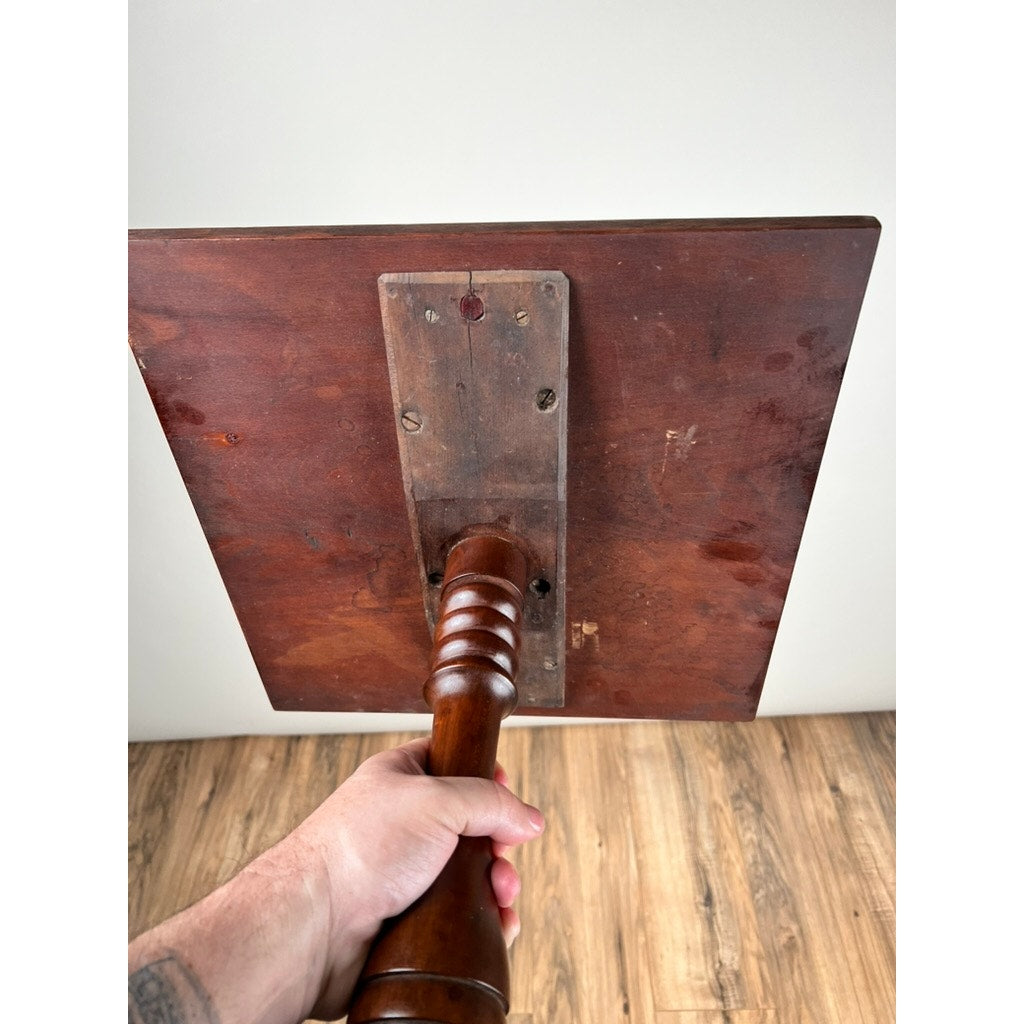 A hand holds an Antique Low Cherry Candle Stand (c. Mid 19th Century) with a square, flat panel, turned handle, and metal plate on the back, shown against a light wall and wood floor. Brand is unknown.