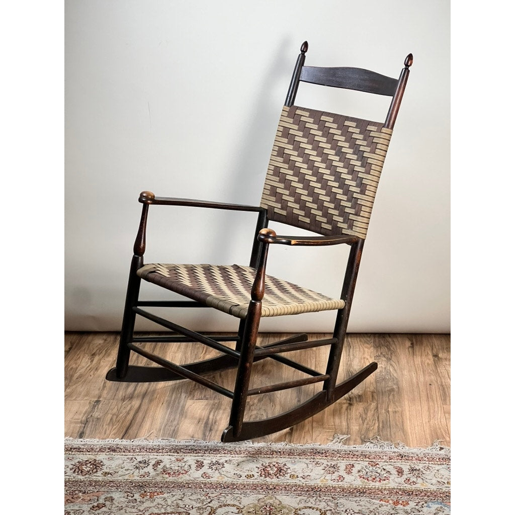 An Antique Shaker #6 Tape Seat Rocker by Mt. Lebanon Shakers, late 19th century, with a woven seat and backrest in tan tape and a dark wood frame, sits on a hardwood floor near a patterned rug. Brand: None/Unknown.