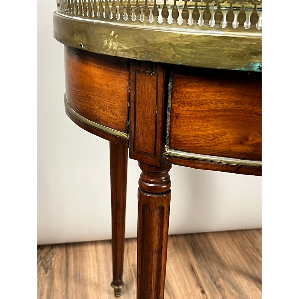 Close-up of an antique 19th-century French Louis XVI-style mahogany bouillotte table with tapered legs, marble top, and pierced brass gallery, set on a wooden floor against a plain wall. Brand unknown.