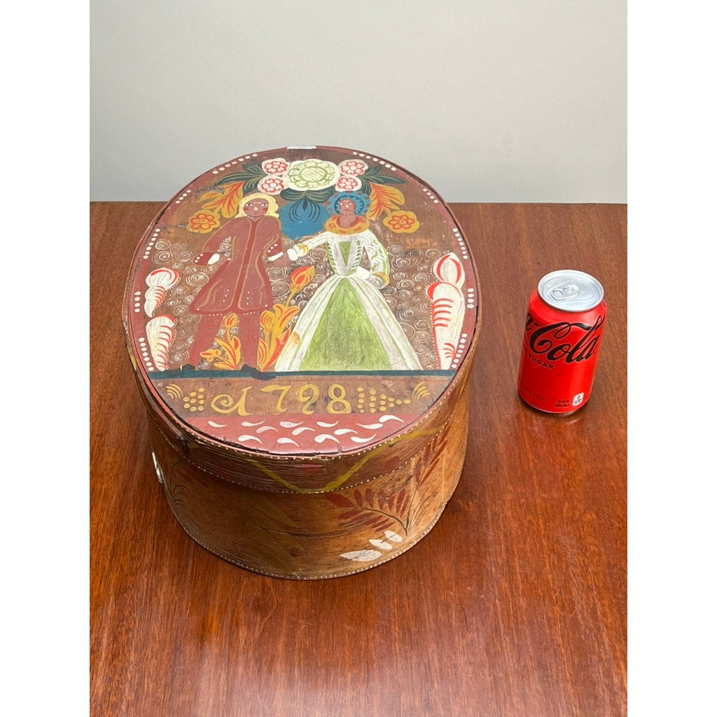 An Antique Swedish Polychrome Painted Marriage Box dated 1798, an oval bentwood piece with traditional folk art decoration, sits on a wooden table next to a red Coca-Cola can, reflecting Scandinavian heritage.