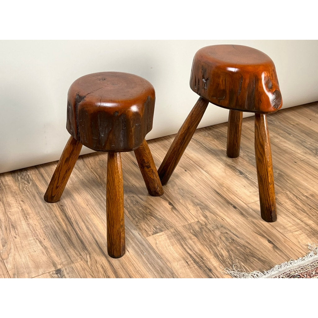 A pair of vintage live edge Wabi Sabi cherry stools signed by Neil Panyik, with three angled legs and natural wood seats, are placed on a wooden floor beside a white wall, embodying the Wabi Sabi aesthetic.