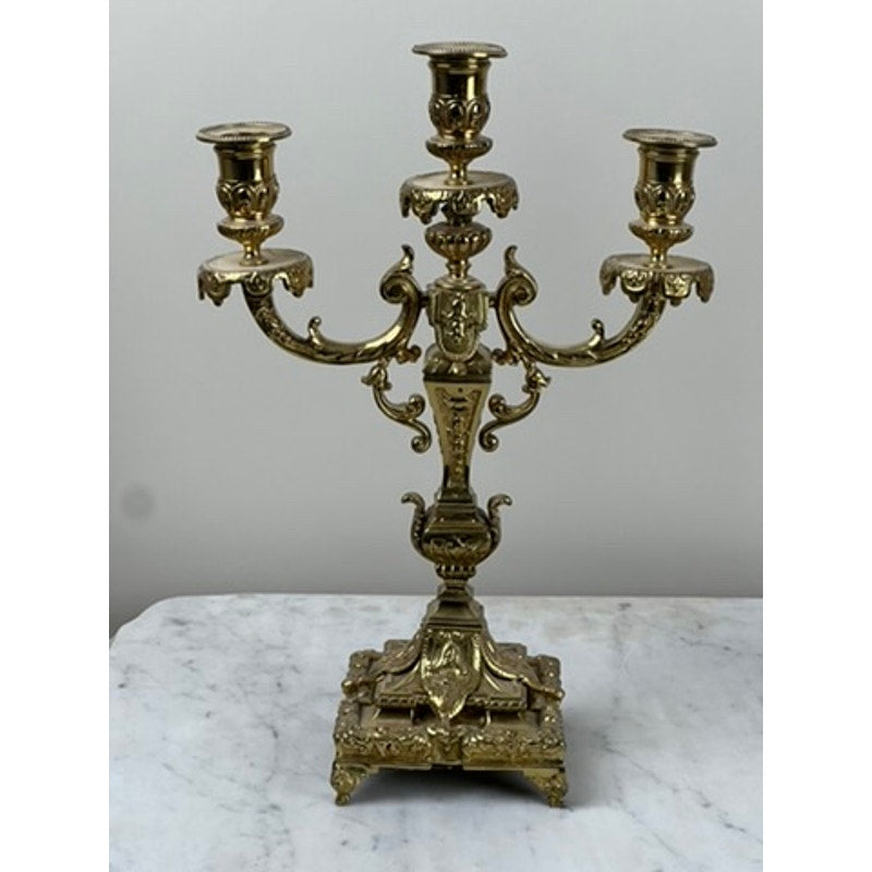 An Antique Pair of late 19th-century brass three-light candelabra, featuring intricate scrollwork and decorative details, is displayed on a white marble surface against a plain light background.