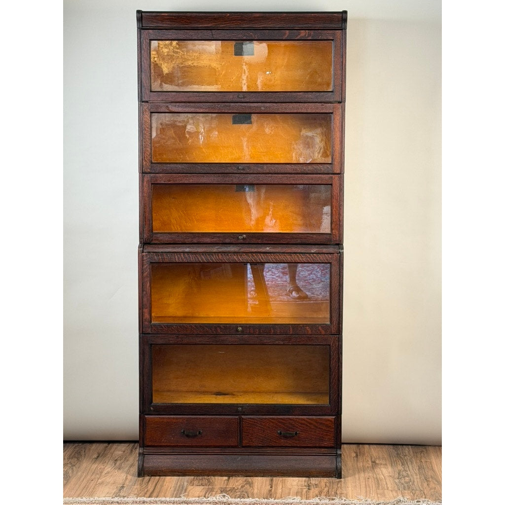 An Antique Hale System Quarter-Sawn Oak Five-Stack Barrister Bookcase, c. 1910 by Camden Cabinet Company, crafted in the Arts and Crafts style with five glass-front stacking shelves and two small drawers at the base, stands gracefully on a wood floor against a plain wall.