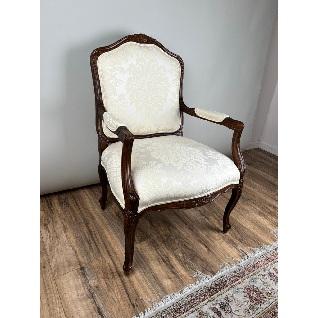 A Vintage Louis XV Style Fauteuil Arm Chair by Pennsylvania House, featuring a carved dark wood frame and cream floral upholstery, sits on a wood floor next to a patterned rug.