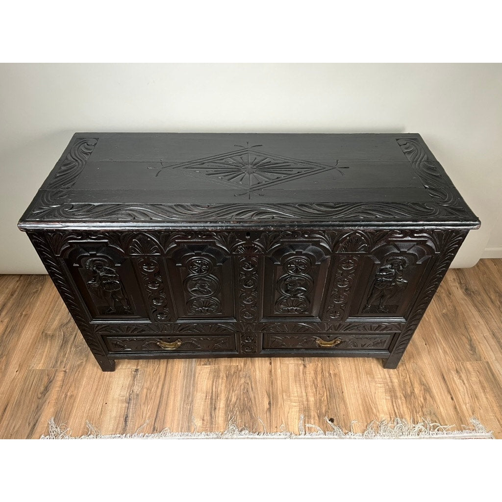 An Antique 18th Century Charles II Style Mule Chest, possibly Irish or Scottish, with carved doors and top plus two brass handles at the base, stands on a wooden floor beside a fringed rug. Brand is unknown.