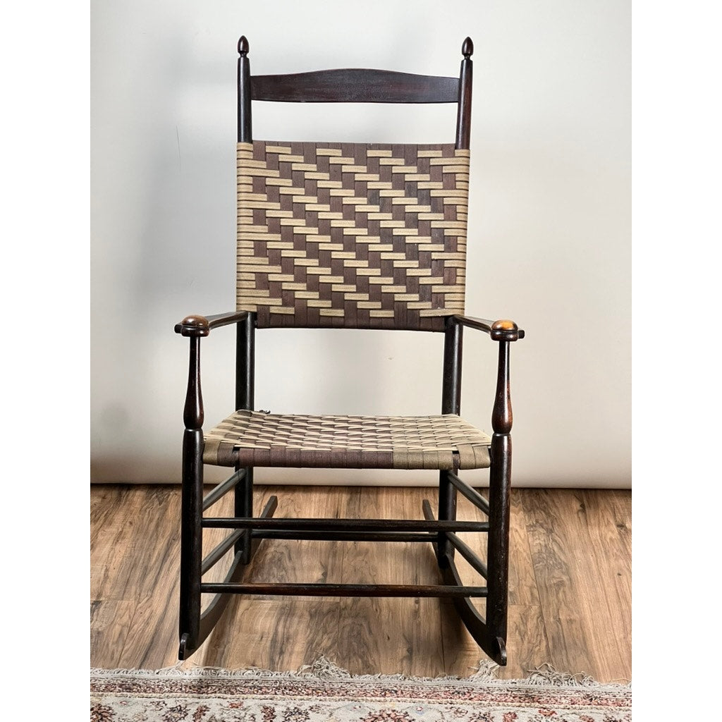 An Antique Shaker #6 Tape Seat Rocker by Mt. Lebanon Shakers, c. late 19th century, with a woven geometric tape seat and backrest, dark wood frame, and armrests sits on a wooden floor near a patterned rug.