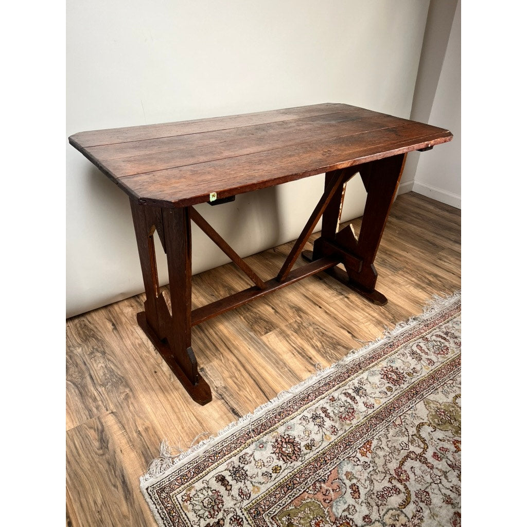 An Antique Rustic Italian Fratino Trestle Table (late 19th century) with a rectangular wood top stands on a wood floor next to a patterned rug, its sturdy trestle legs adding timeless farmhouse charm against the plain wall.