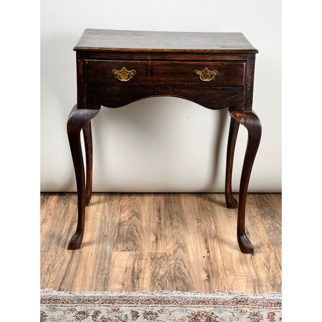 An Antique Queen Anne Style George III Oak One Drawer Stand (late 18th century) with cabriole legs and brass handle sits on a wood floor, with part of a patterned rug visible at the bottom. Brand: None/Unknown.