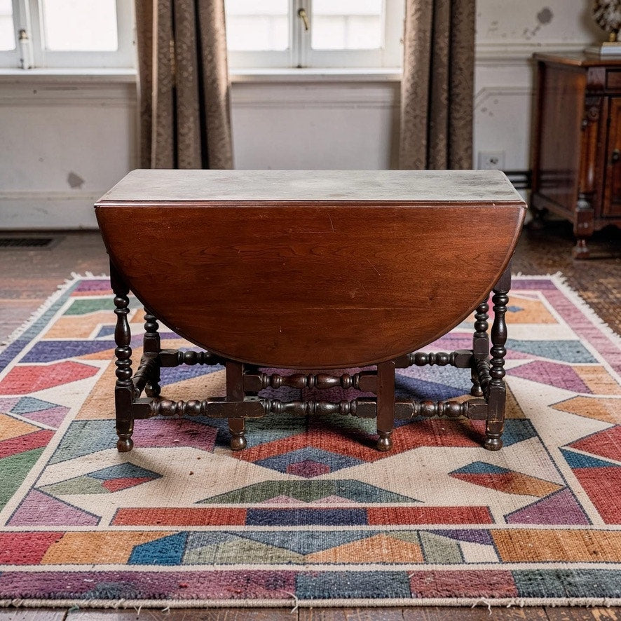 An Antique English William & Mary Walnut One Drawer Gateleg Dining Table (early 18th century) stands on a colorful geometric rug in a light-walled room with curtains and a window in the background.