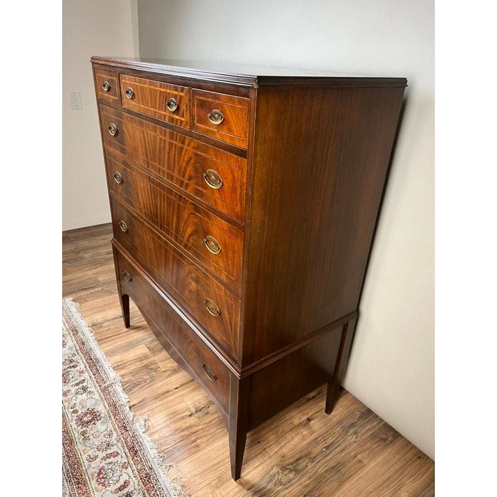 A tall, vintage Dutch Woodcraft Shops Federal Style Flame Mahogany Chest on Stand (c. 1945–1955) with six drawers, brass handles, and tapered legs stands on a wooden floor beside a patterned rug and a white wall. This striking piece showcases the renowned craftsmanship of Dutch Woodcraft Shops, making it a unique accent in any room.