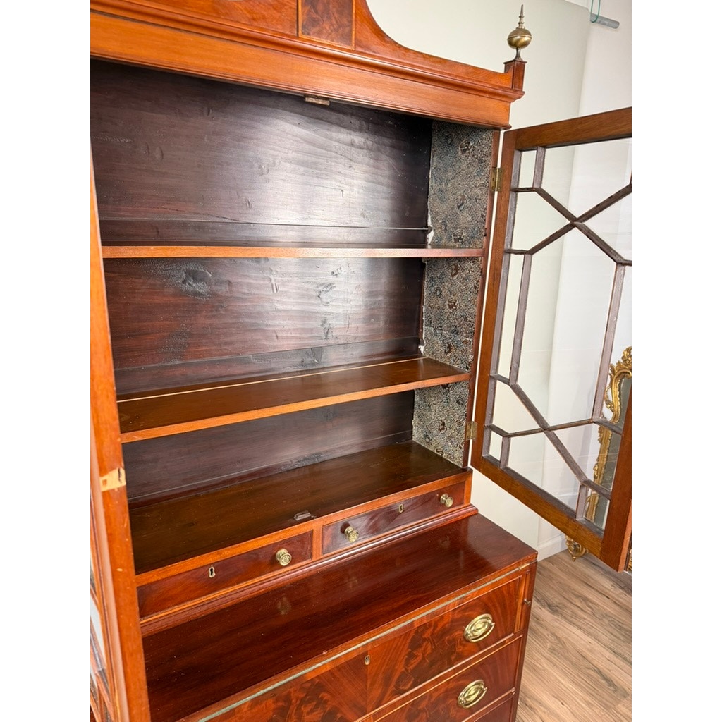 Federal Flame Mahogany Secretary Bookcase with Eagle Finial, c. 1810–1825. This open wooden cabinet features a glass door, shelves, and drawers, with notable dark mold or water damage on the right interior side. Crafted in the tradition of early 19th-century American cabinetmaking, it is fitted with brass handles and stands on a wooden floor near a gold-framed mirror. Brand: Unknown.