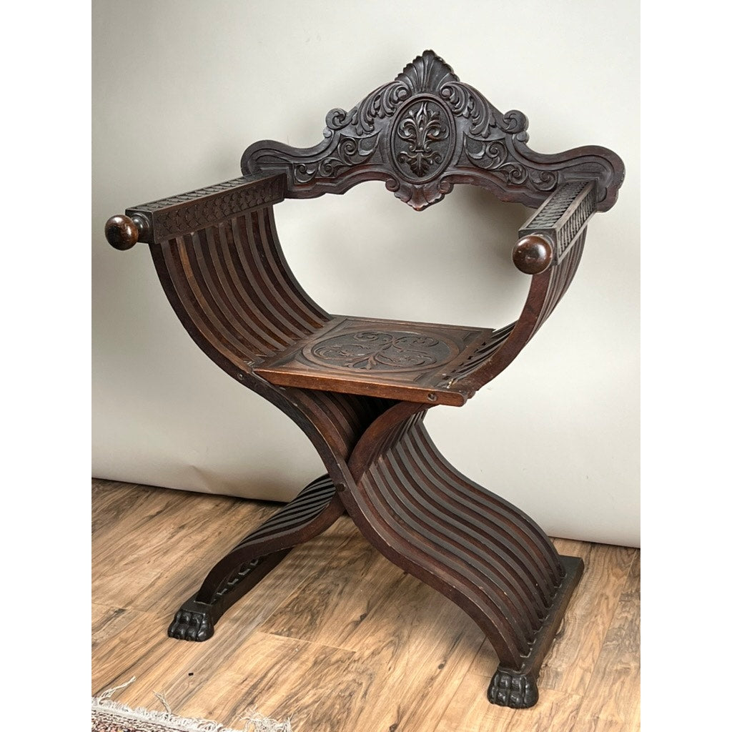 Antique Italian Renaissance Walnut Savonarola Chair, late 19th century, with curved X-shaped legs, intricately carved backrest, decorative seat, arms ending in rounded knobs, and claw feet; set on a wooden floor against a plain background.