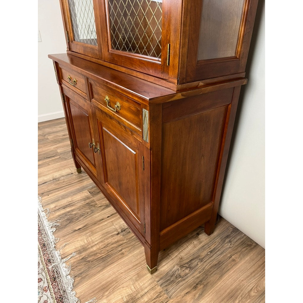 The Vintage William IV Style Cherry Glass Door Bookcase by Stickley features diamond-patterned glass doors, brass handles, two drawers, and lower storage. This elegant Stickley piece stands on a wooden floor near a cream wall and patterned rug.