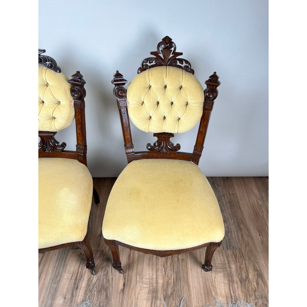 A Fine Antique Pair Renaissance Revival Walnut Side Chairs, attributed to John Jelliff (Newark, c. 1880), with carved details and yellow tufted upholstery, sit on a wooden floor by a light gray wall; only part of the second chair is visible.