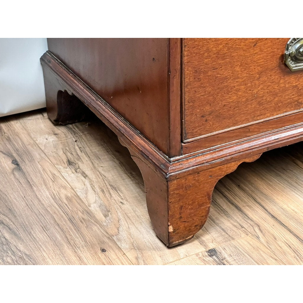 Close-up of the corner of a Late Georgian English Mahogany Chest of Drawers, circa 1780–1810, highlighting its decorative bracket feet as it rests gracefully on a wood floor. This period piece displays a natural finish with prominent wood grain and includes a partially visible brass handle.