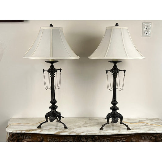 A Vintage Pair of French Empire Revival Table Lamps with Oil Rubbed Bronze Finish by Pearl City Antiques, each topped with a white fabric shade, are displayed on a marble-topped antique wooden table in front of a light-colored wall featuring a visible power outlet above.