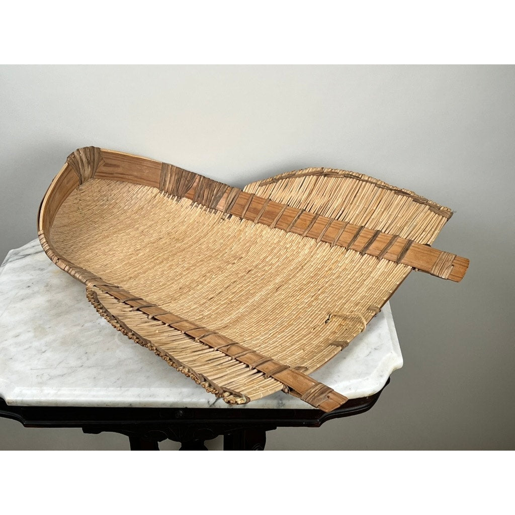 The Vintage Traditional Winnowing Basket, made from natural fibers with visible weaving patterns and raised edges, sits on a white marble-topped table, adding rustic farmhouse charm with its large, curved shape. Brand: Unknown.