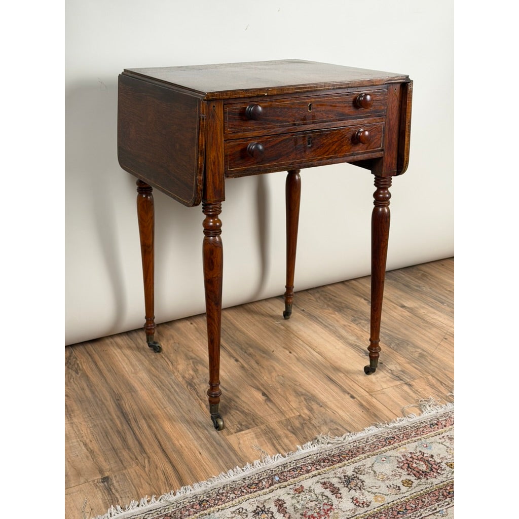 An English Regency Rosewood Two-Drawer Drop-Leaf Work Table with Brass and Satinwood Inlay, circa 1820–1840, featuring two stacked drawers and slender, turned legs on casters.