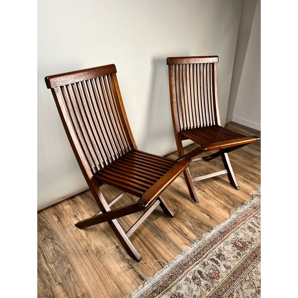 A Pair of Vintage Mid-Century Modern Style Teak Folding Chairs with slatted backs and seats, featuring a rich dark brown finish, are placed on a wooden floor next to a patterned rug and a plain light-colored wall. Brand unknown.