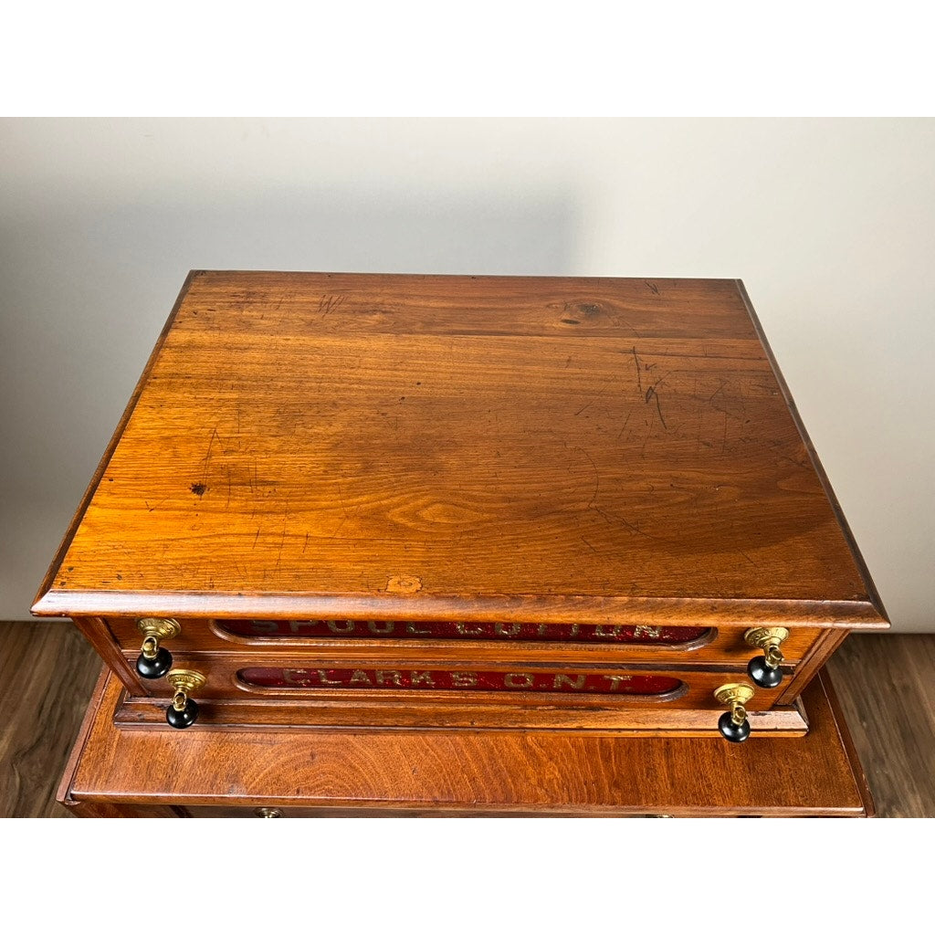 The Antique Victorian Pine Two Drawer Clark’s O.N.T Spool Cabinet by Clark's ONT, c. late 19th century, features brass handles and red labels on each drawer, a polished, slightly scratched finish, and rests on a wooden floor against a light wall.