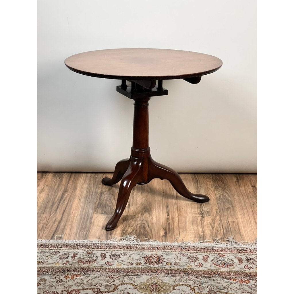 An Antique Queen Anne Mahogany Birdcage Tilt Top Breakfast Table (early 19th century) with a tripod base stands on a wooden floor beside a patterned rug, set against a plain white wall.