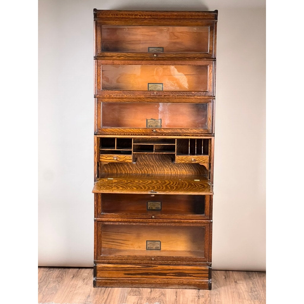 An Antique Globe-Wernicke Quarter Sawn Oak 6-Stack Barrister Bookcase Secretary Desk (c. 1905–1915) by Globe Wernicke stands on a wood floor against a plain light-colored wall, featuring six glass-fronted sections including a midsection drop-down secretary desk with small pigeonhole compartments.