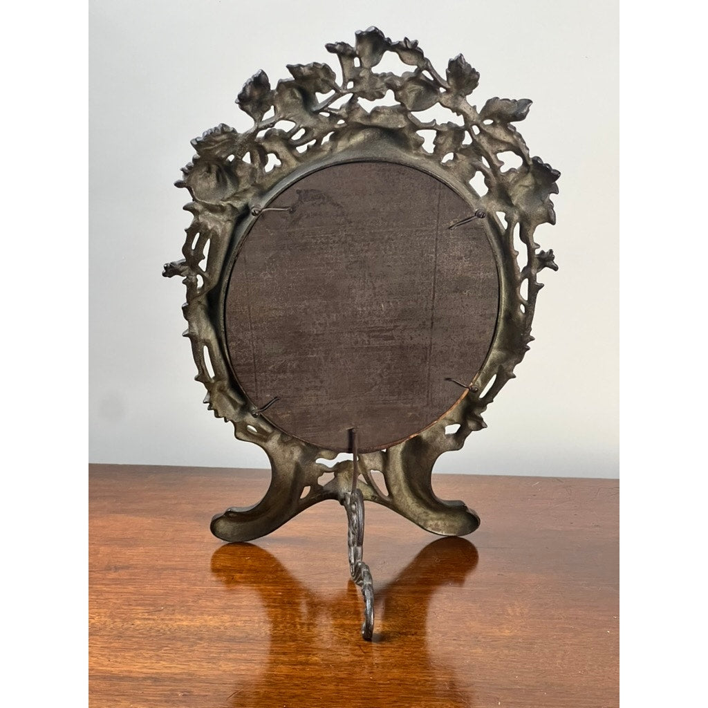 An Antique Art Nouveau Silvered Cast Brass Rose Vanity Dressing Mirror, dated 1909, stands on a wooden surface with its ornate floral back and stand visible against a plain light background. Brand: Unknown.
