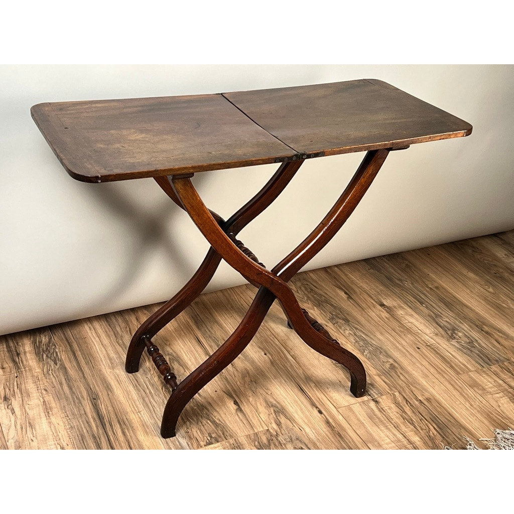 An Antique English Regency Mahogany Folding Coaching or Sporting Table (c. 1825–1845) with a rectangular top and two curved intersecting legs stands on a wooden floor against a plain light-colored wall.