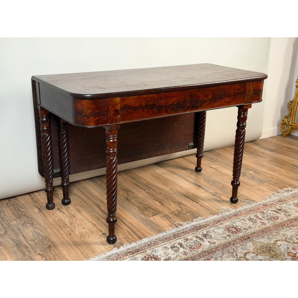 The Pearl City Antiques American Classical Mahogany Single Drop-Leaf Table with Spiral-Twist Legs, c. 1830–1845, showcases four elegant spiral-twist legs, a rich dark wood finish, and a single drop-leaf design with a fold-out top. It is pictured on a wooden floor beside a patterned rug and against a white wall.