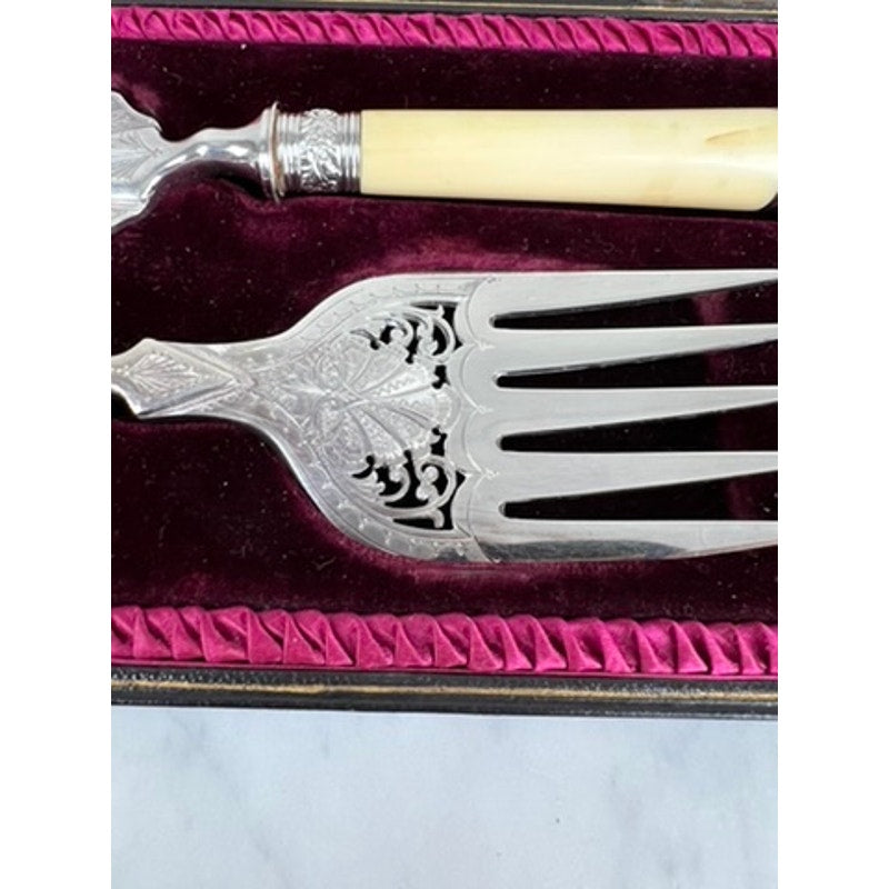 A close-up of an Antique Victorian Silver-Plated Fish Serving Set, featuring a serving knife and fork with ornate handles and intricate cut-out designs, displayed in its original velvet-lined presentation box with a deep red border.