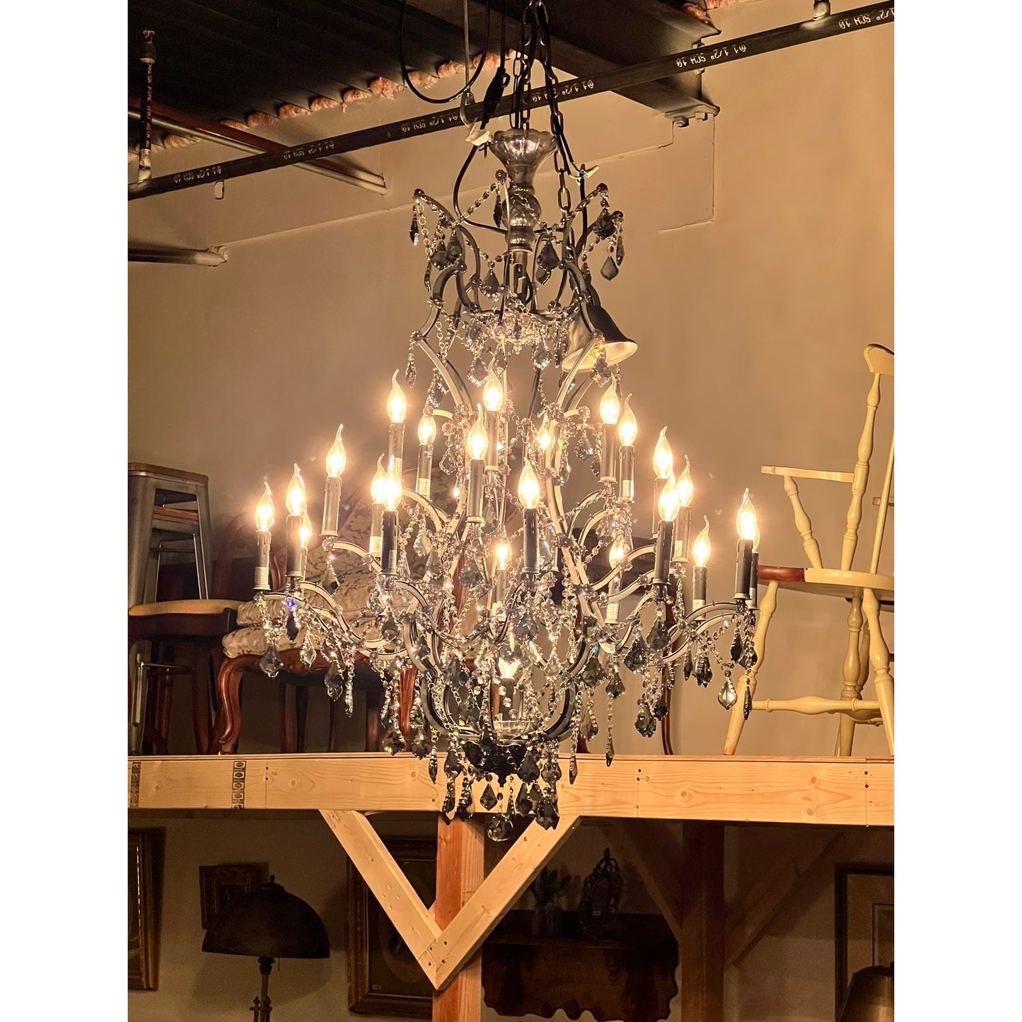 A Large Vintage Smoke Crystal 24 Light Chandelier hangs from the ceiling of a room with wooden beams, stacked chairs, and framed pictures on the walls in the background.