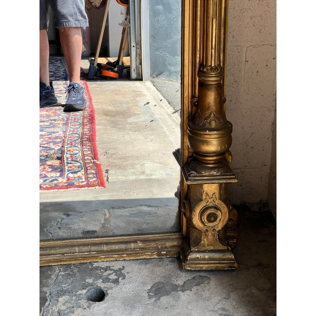 The bottom corner of an Exceptional Monumental Victorian Renaissance Revival Pier Mirror, circa 1870–1885, with an ornate giltwood frame rests on a concrete floor, reflecting part of a rug and a person wearing gray shorts and sneakers.