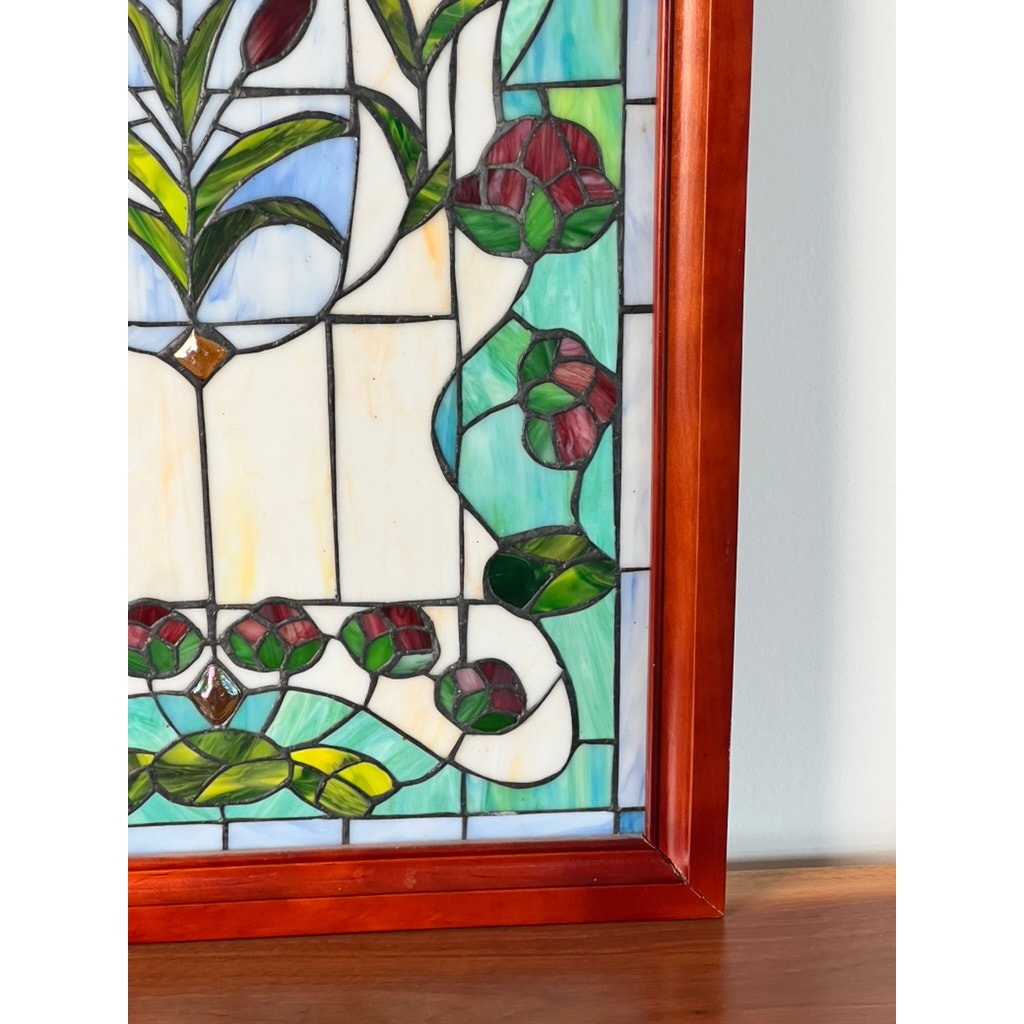 A close-up of the Vintage Stained Glass Wall Hanging 32x20 with a wooden frame, displaying red flowers, green leaves, and blue-and-white sections. This decorative piece rests on wood against a light-colored wall. Brand: None/Unknown.