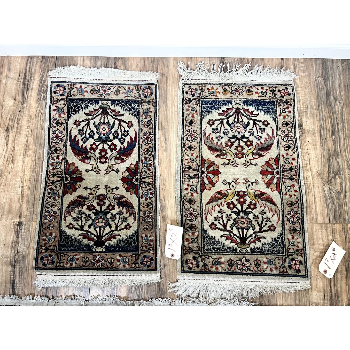 A vintage pair of hand-knotted Oriental Tree of Life scatter rugs (1.5 x 2.5), each with intricate floral and bird motifs, white fringe, and price tags attached, lie side by side on a wood floor. Brand: None/Unknown.